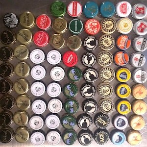 May include: A collection of colorful bottle caps with various brand names and logos, including Guinness, Stella Artois, Beck's, Angry Orchard, and London Pride. The caps are arranged in a grid pattern on a silver surface.