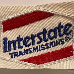 May include: Embroidered patch with the words "Interstate Transmissions" in blue, red, and white. The patch has a white background and a red and white border. The text is in a bold, stylized font.