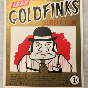 Może przedstawiać: Karta gumy do żucia Goldfinks w stylu vintage z kreskówkowym mężczyzną w meloniku i koszuli w paski. Karta ma złote tło z napisem "GOLDFINKS BUBBLE GUM" i "LEAF". Karta zawiera również tekst "1¢".