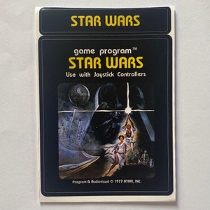 May include: A vintage Star Wars game program label. The label features the Star Wars logo in yellow at the top and the game title in yellow. The artwork depicts Darth Vader, Luke Skywalker, Princess Leia, and the Death Star. The text reads "Use with Joystick Controllers."