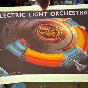Może przedstawiać: Plakat vintage dla Electric Light Orchestra. Plakat przedstawia projekt statku kosmicznego z logo zespołu pośrodku. Statek kosmiczny ma niebieskie, pomarańczowe i żółte akcenty. Tekst "ELECTRIC LIGHT ORCHESTRA" znajduje się u góry na różowo.