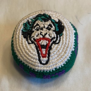 May include: A round, crocheted item featuring an embroidered image of the Joker's face. The design includes green hair, a red mouth, and black outlines on a white background. The edges are green and purple.