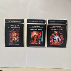 May include: Three game program cards for the Star Wars video game series. The cards are white with black borders and feature the titles "The Phantom Menace", "Attack of the Clones", and "Revenge of the Sith". Each card has a colorful image of characters from the corresponding movie.