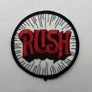 May include: Embroidered patch with the word "RUSH" in red, stylized lettering. The patch is circular, with a black border and a white background featuring radiating lines. The text is set against a black background.