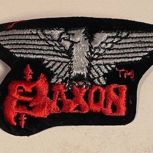May include: A black embroidered patch featuring a red "SAXON" logo and a silver eagle with outstretched wings. The eagle is positioned centrally above the logo, with the text "TM" to the right of the logo.