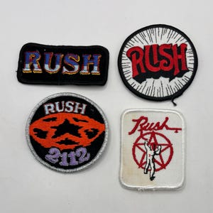 May include: Four embroidered patches featuring the band name Rush. One rectangular black patch with blue and yellow text, one circular white patch with red text, one circular black patch with an orange star and 2112, and one white square patch with a red star.