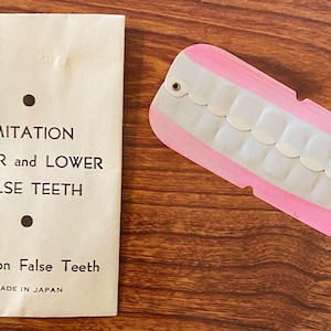 May include: A package of imitation upper and lower false teeth, made in Japan. The package is white with black text and the teeth are pink and white.