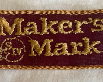 May include: A burgundy fabric patch with gold embroidered text that reads "Maker's Mark" and a gold embroidered star and circle with the letters "SM" inside.