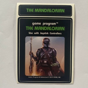 May include: A black and green game cartridge with a picture of a character from the Star Wars series The Mandalorian. The text on the cartridge reads "game program™ THE MANDALORIAN Use with Joystick Controllers Label, Program & Audiovisual © 2019 ATARI, INC."