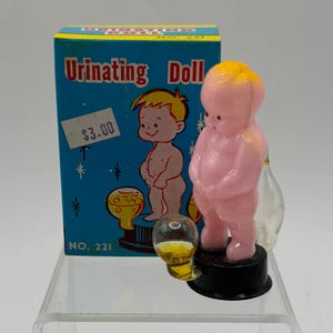 Könnte beinhalten: Ein Vintage-Novelty-Spielzeug, eine rosa Plastikpuppe mit gelben Haaren, die auf einem schwarzen Sockel steht. Die Puppe befindet sich vor einer blauen Schachtel mit der Aufschrift "Urinating Doll" und einer Cartoon-Illustration.