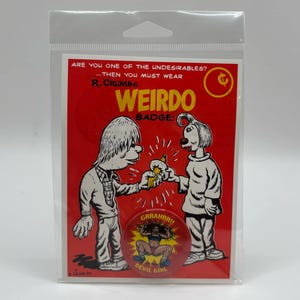 Pode incluir: Uma impressão gráfica vermelha e branca com duas figuras de desenho animado e o texto "WEIRDO BADGE!". Um alfinete circular com o texto "GRRAHRR!! DEVIL GIRL" também é visível. A imagem está em uma manga de plástico transparente.