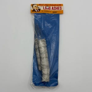 Pode incluir: Um item de novidade embalado em um saco plástico transparente, apresentando dois objetos cilíndricos longos que se assemelham a cinzas de cigarro. A embalagem inclui um fundo azul e um cartão de cabeçalho com o texto "Feel your friends with LONG ASHES" e uma ilustração de desenho animado.