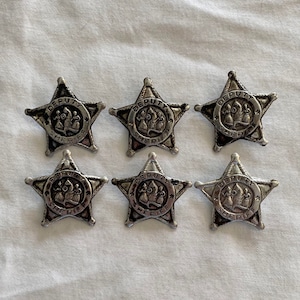 Puede incluir: Seis insignias de estrella de sheriff plateadas con las palabras "DEPUTY SHERIFF" alrededor del borde. Cada insignia presenta una imagen estilizada de un sombrero de sheriff y una insignia.