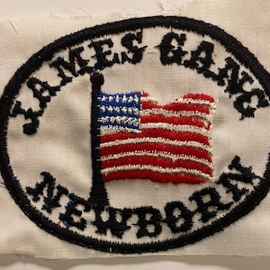 Può includere: Una toppa di stoffa bianca con un cerchio nero attorno al bordo. Il cerchio ha il testo "JAMES GANG NEWBORN" in lettere nere. All'interno del cerchio c'è una bandiera americana rossa, bianca e blu.