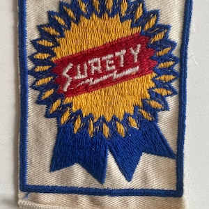 Op de afbeelding: Een vintage geborduurde patch met een blauw lint en een gele ster. De ster heeft een rood banner met het woord "SURETY" in witte letters.