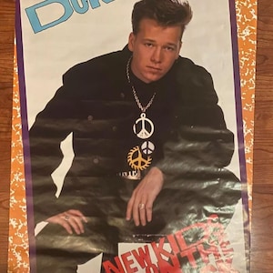 Op de afbeelding: Een vintage poster met Donnie Wahlberg van New Kids on the Block. De poster is oranje en paars met een witte achtergrond. Donnie draagt een zwart jack met een zilveren ketting met vredesemblemen. De tekst "DONNIE" is blauw en de tekst "NEW KIDS ON THE BLOCK" is rood.