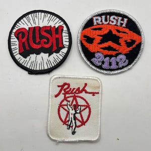 May include: Three embroidered patches featuring the band Rush. One circular patch has a white background with black lines and red text. Another circular patch is black with an orange star and purple 2112 text. A square patch shows a red star and figure.