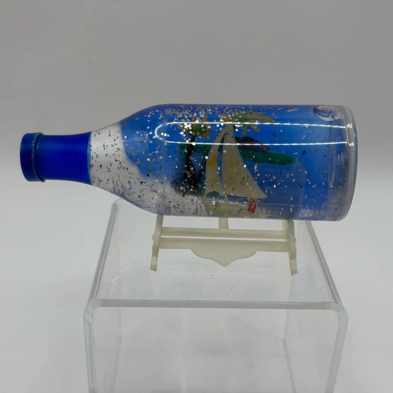 May include: A clear, bottle-shaped snow globe with a blue neck and a tropical scene inside. The scene includes a sailboat, palm trees, and silver glitter suspended in blue liquid. The globe is displayed on a white plastic stand.