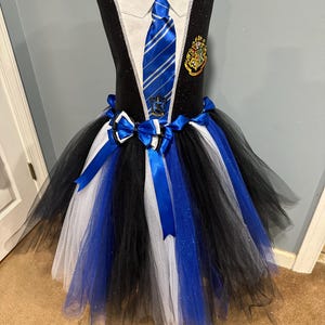 May include: A black, white, and blue tutu dress with a school uniform-inspired bodice. The bodice features a white collar, a blue striped tie, and a school crest. The tutu skirt is layered with black, white, and blue tulle, and adorned with blue bows.