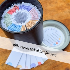 May include: A black cylindrical container filled with colorful cards, with the text "100+ Verses picked just for you!". The cards are arranged in a circular pattern. A lid with a color-coded feeling chart is next to the container.