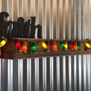 May include: A wooden shelf with a metal sign that says "faith" above it. The shelf is decorated with a string of lights made from shotgun shells. The shells are painted in various colors, including green, yellow, red, and orange.