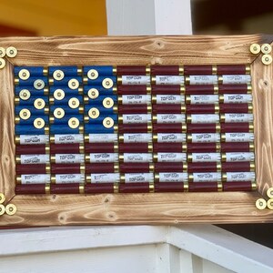 May include: A wooden framed American flag made of shotgun shells. The shells are arranged in the pattern of the American flag with red, white, and blue colors. The shells are labeled "Top Gun" with a white label on each shell.