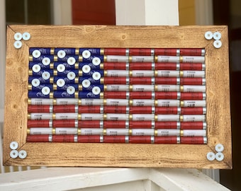 May include: A rectangular wall art piece featuring a flag design constructed from blue, red, and white shotgun shells arranged in a grid pattern. The assembly is encased in a rustic, light brown wooden frame with white shell bases at each corner.