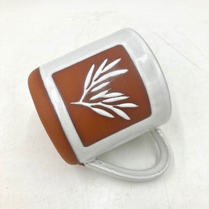 Può includere: Tazza in ceramica bianca con un design quadrato marrone che presenta un ramo stilizzato bianco. La tazza ha un manico bianco.