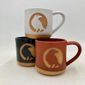 May include: Three ceramic mugs stacked together, each featuring a circular cutout design of a crow. The top mug is white, the bottom left is black, and the bottom right is terracotta orange. Each mug has a natural, unglazed tan base and handle.