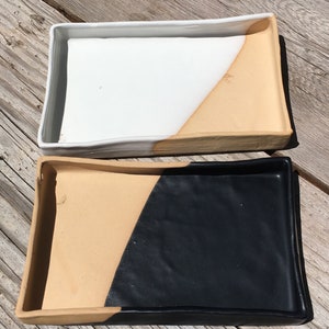 Può includere: Due vassoi rettangolari in ceramica con un design unico a blocchi di colore. Il vassoio superiore è bianco con un bordo color sabbia, mentre il vassoio inferiore è color sabbia con un bordo nero.