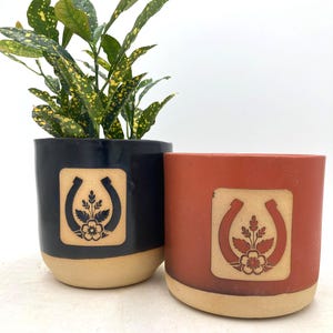 Könnte beinhalten: Zwei Keramik-Pflanztöpfe, einer schwarz und einer rotbraun, mit beigem Sockel. Jeder Topf hat ein quadratisches Design mit Hufeisen- und Blumenmotiv. Eine Pflanze mit grünen und gelben Blättern befindet sich im schwarzen Topf.