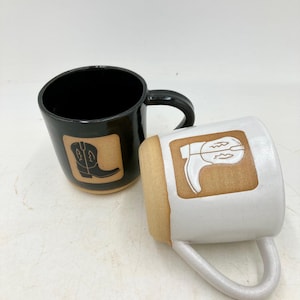 Può includere: Due tazze in ceramica dal design rustico. Una tazza è nera con un disegno di stivale da cowboy, l'altra è bianca con un disegno di stivale da cowboy. Entrambe le tazze hanno uno sfondo quadrato marrone e un manico curvo.