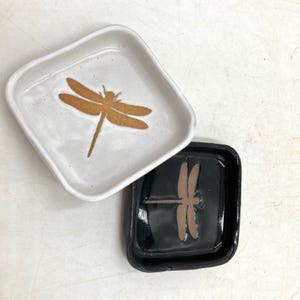 Può includere: Due piatti quadrati in ceramica con disegni di libellule. Un piatto è bianco con una libellula dorata, e l'altro è nero con una libellula marrone.