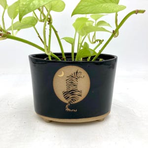 以下が含まれることがあります： 黒いセラミック植木鉢。茶色のベースと、虎のシルエットと三日月が描かれた円形デザイン。緑の葉の植物が入っています。光沢のある仕上がりで、小さな足で持ち上げられています。