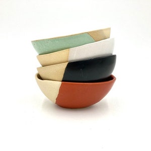 Può includere: Una pila di cinque piccole ciotole in ceramica di vari colori. Le ciotole sono nelle tonalità di verde, bianco, nero e arancione, con un accento beige naturale su ciascuna. Le ciotole sono impilate l'una sull'altra.