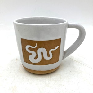 Puede incluir: Una taza de cerámica blanca con un asa gruesa y un parche rectangular marrón en la parte frontal. El parche marrón presenta un gráfico blanco de una serpiente sinuosa y estilizada. La base de la taza es de arcilla marrón sin esmaltar.