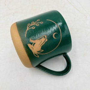 May include: A ceramic mug with a forest green exterior and a tan-colored rim and interior. The mug features a tan-colored illustration of a wolf, crescent moon, and botanical elements.