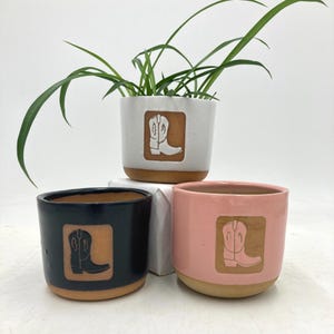 Puede incluir: Tres macetas de cerámica en blanco, azul marino y rosa, cada una con un diseño de bota de vaquero. Las macetas tienen una base de arcilla natural y contienen vegetación. El diseño de la bota de vaquero está en un cuadrado.