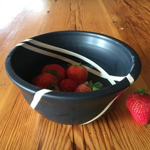 Può includere: Una ciotola nera con strisce bianche, piena di fragole rosse. Una fragola si trova fuori dalla ciotola su una superficie di legno.