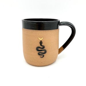May include: A tan ceramic mug with a black rim and handle. The mug has a black snake design with a gold crescent moon above it.