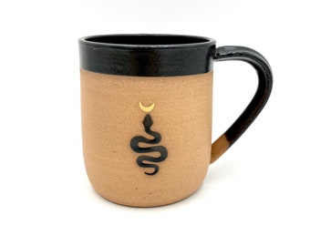 May include: A tan ceramic mug with a black rim and handle. The mug has a black snake design with a gold crescent moon above it.