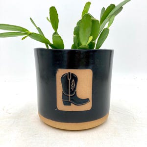 Puede incluir: Una maceta de cerámica negra con base marrón y un cuadrado marrón con un diseño de bota de vaquero negra. Contiene una planta suculenta verde. El fondo es blanco.