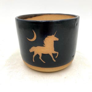 May include: A cylindrical ceramic mug with a matte black exterior and a natural tan clay base. The front features a carved silhouette of a unicorn and a crescent moon, revealing the tan clay color beneath the black glaze on the surface.