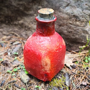 May include: A small, red, ceramic bottle with a cork stopper. The bottle has a rough, textured surface and is sitting on a bed of pine needles and moss.