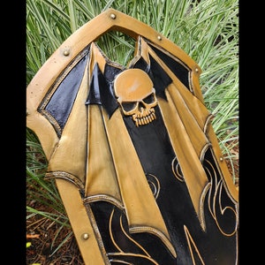 K&ouml;nnte beinhalten: Ein schwarzer und goldener Schild mit einem Totenkopfdesign. Der Schild ist mit komplizierten Details verziert und hat einen erh&ouml;hten Rand.