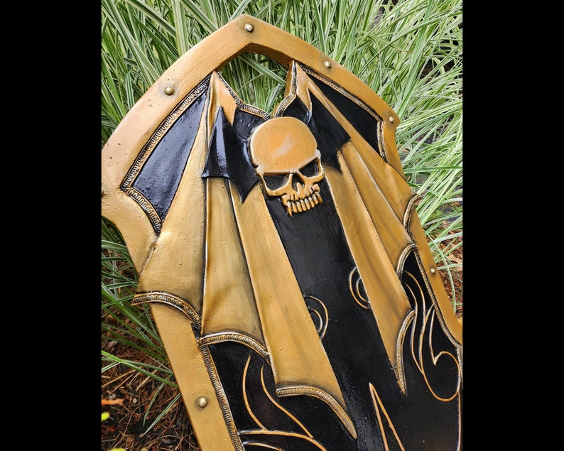 K&ouml;nnte beinhalten: Ein schwarzer und goldener Schild mit einem Totenkopfdesign. Der Schild ist mit komplizierten Details verziert und hat einen erh&ouml;hten Rand.