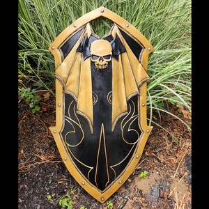 K&ouml;nnte beinhalten: Ein schwarzer und goldener Schild mit einem Totenkopf- und Fl&uuml;geldesign. Der Schild ist aus Holz und hat eine abgenutzte Oberfl&auml;che.