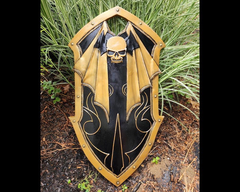 K&ouml;nnte beinhalten: Ein schwarzer und goldener Schild mit einem Totenkopf- und Fl&uuml;geldesign. Der Schild ist aus Holz und hat eine abgenutzte Oberfl&auml;che.