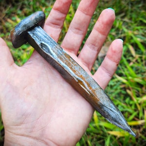 May include: A large, rusty, hand-forged nail with a square head. The nail is about 8 inches long and has a pointed tip.