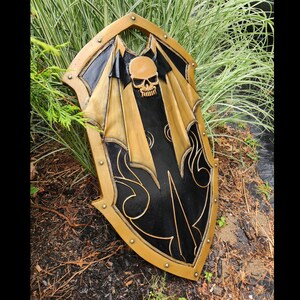 K&ouml;nnte beinhalten: Ein schwarzer und goldener Schild mit einem Totenkopf- und Fl&uuml;gelmotiv. Der Schild ist aus Holz und hat eine abgenutzte Oberfl&auml;che.